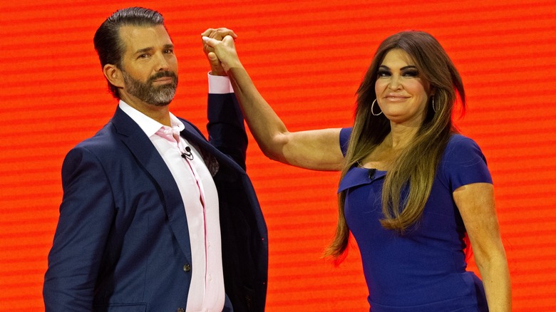 Donald Trump Jr. and Kimberly Guilfoyle holding raised hands red background