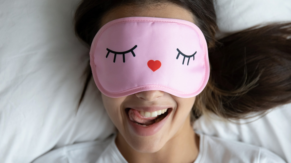 Woman wearing a pink sleep mask while lying on a white pillow and smiling.