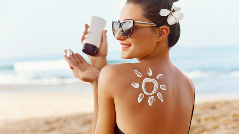 Woman on a beach wearing a black bikini and a sunhat applying sunscreen.