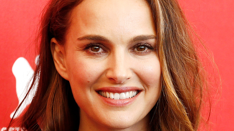 Natalie Portman poses with a big smile in front of a red background.