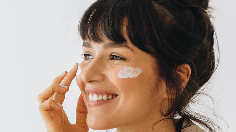 A woman smiling while applying moisturizer on her face.
