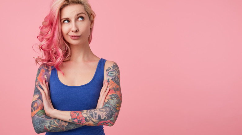 Woman with pink and blonde hair and blue tank top with tattooed arms folded is looking up with a questioning look